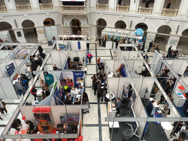Große Gruppe von Besuchern auf einer internationalen Fachmesse mit Ständen, Bannern, Tischen, Stühlen und Ballons in einem Saal mit gewölbter Architektur und einem Bildschirm im Hintergrund.