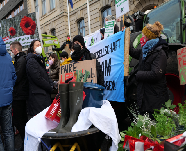 Maskierte Demonstranten mit Protestschildern vor einem Lkw mit einem Tisch, Pflanzen und einem Gebäude mit Flagge im Hintergrund.