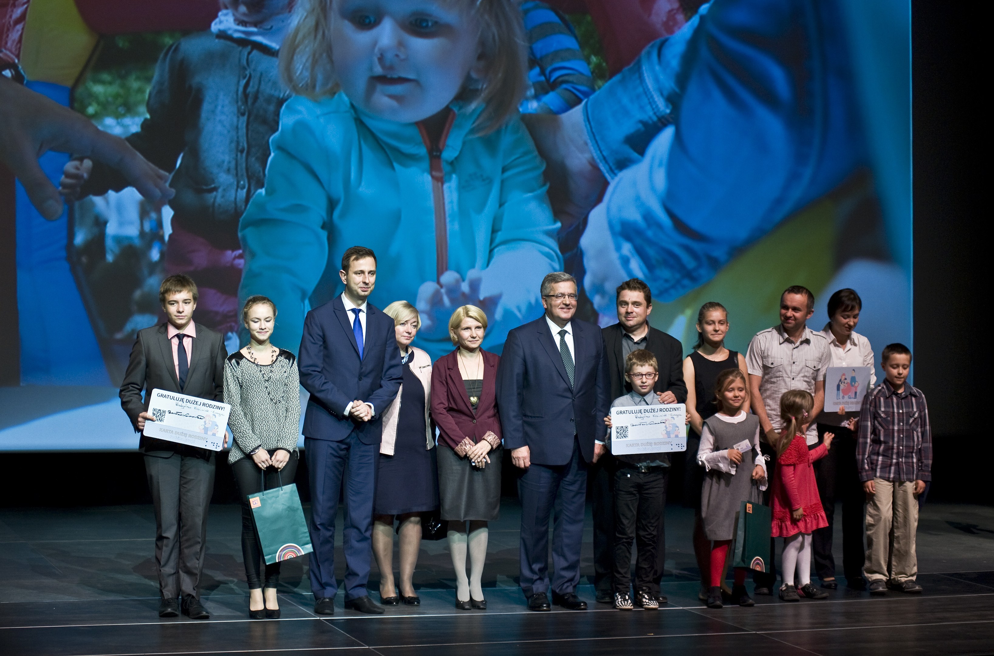 Gruppe von Menschen auf einer Bühne mit Taschen und Urkunden, mit einem Bildschirm, der ein Bild eines Kindes und andere im Hintergrund anzeigt.