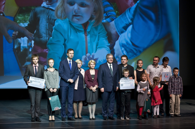 Gruppe von Menschen auf einer Bühne mit Taschen und Urkunden, mit einem Bildschirm, der ein Bild eines Kindes und andere im Hintergrund anzeigt.