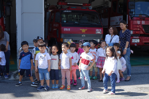 Eine Gruppe von Kindern vor einem Feuerwehrauto an einer Feuerwache, einige tragen Mötzen, mit weiteren Feuerwehrautos im Hintergrund.