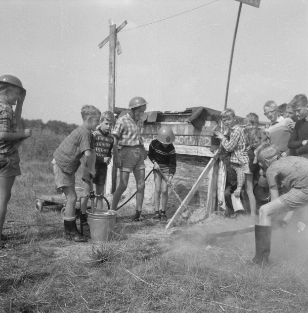 Schwarz-weißes Bild von Kindern in Helmen um ein Hydrant in einem grasbewachsenen Feld versammelt, mit einem Eimer, einem Karren, Stangen mit Drähten und einer Texttafel in der Nähe, Bäume im Hintergrund unter einem klaren Himmel.