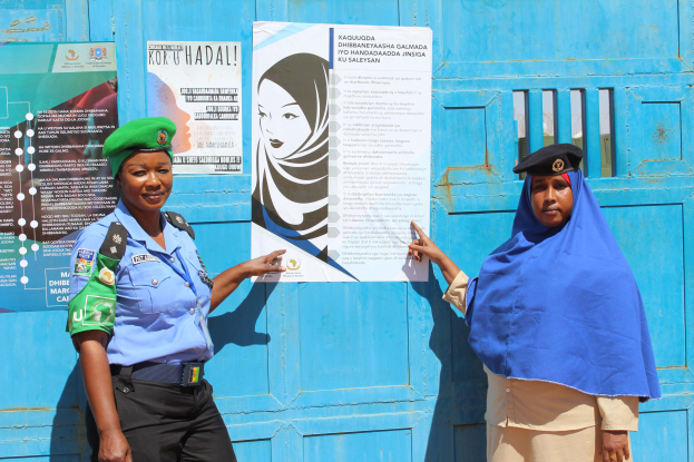 Zwei Frauen stehen nebeneinander vor einer blauen Tür, die mit Plakaten bedeckt ist. Eine trägt ein blaues Hemd, schwarze Hosen und eine grüne Mütze, die andere hält ein Papier in der Hand.