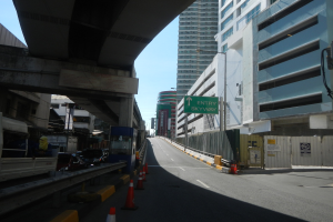 Stadtstraße mit Verkehrskegeln, Fahrzeugen, einer Brücke auf der linken Seite, Gebäuden mit Namensschildern, Laternenmasten und einem Himmel als Hintergrund.