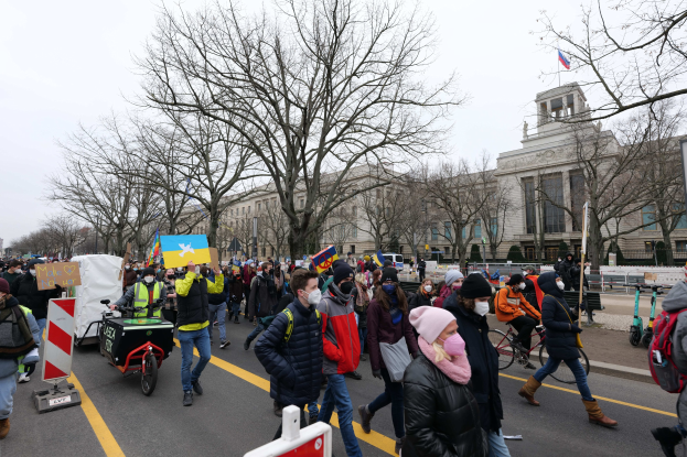 Ein großer Protestmarsch mit Menschen, die eine Straße in Washington, D.C. entlanggehen, einige halten Schilder und andere fahren Fahrräder, mit Bäumen, Schildern und einem klaren blauen Himmel im Hintergrund.