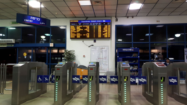 Ein Mann steht vor einer Metallschranke am Flughafen, mit Metalldetektoren, Hinweistafeln, einem Topf mit Pflanze, einer Wandtafel, einer Decke mit Leuchten und einem Anzeigebildschirm.