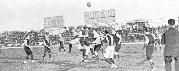 Ein Schwarz-Weiß-Bild einer Gruppe von Männern, die auf einem Feld Fußball spielen, mit einem Ball in der Luft und einer Zuschauermenge im Hintergrund, mit Schildern, die wahrscheinlich textbezogen zur Partie sind.