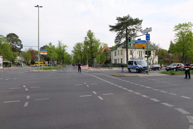 Ein Kreisverkehr mit einem Polizeiwagen am Straßenrand, fahrende Fahrzeuge, Fußgänger auf dem Gehweg, Verkehrsampeln, Bäume, Gebäude mit Fenstern und eine bewölkte Himmel.