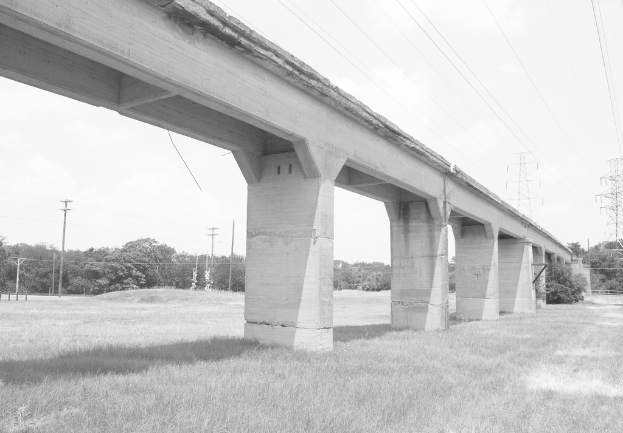 Eine Betonbrücke auf einer grünen Wiese mit einem großen Strommast und Kabeln darüber, umgeben von Bäumen und zusätzlichen Strommasten im Hintergrund.