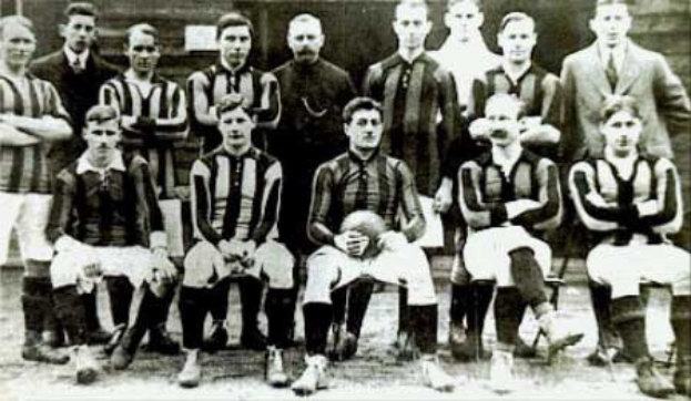 Schwarzes und weisses Foto einer Fussballmannschaft, einige stehen und einige sitzen, vor einer Wand mit dem Text "Ballymena Football Club" unten.