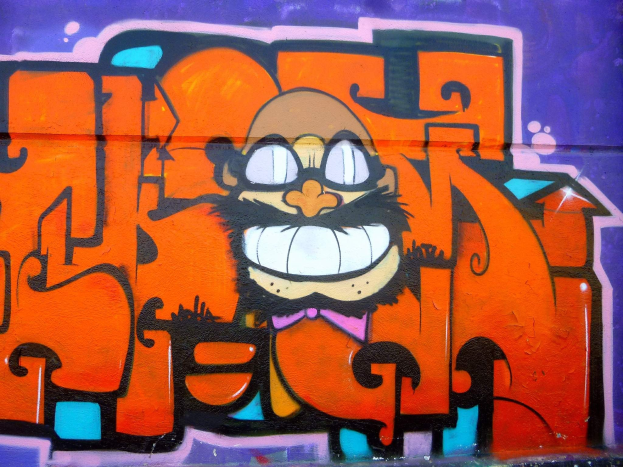 Graffiti-Malerei eines Cartoon-Charakters mit großen Augen, einem breiten Grinsen und einem verschmitzten Gesichtsausdruck an der Seite eines Gebäudes, mit leuchtenden Farben und einer markanten Begleittext.