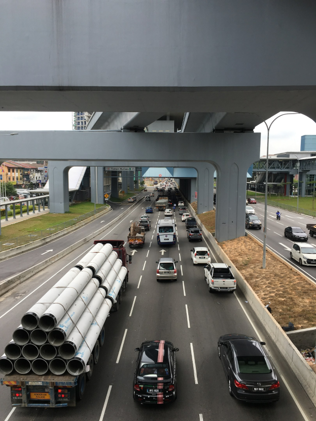 Eine befahrene Autobahn mit mehreren Fahrzeugen, eine Brücke darüber, Straßenlaternen, Gras, Gebäude, Bäume und einen klaren Himmel.