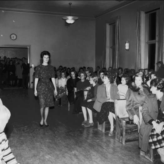 Eine Frau in einem schwarzen und weißen Kleid läuft über einen Laufsteg, während ein Publikum zusieht, bei einer Modenschau, die das Frauenwahlrecht feiert.