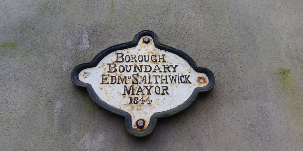 Schwarze Metalltafel an einem Gebäude mit weißer fettgedruckter Schrift, die "Gemeindegrenze Edm Smithwick Bürgermeister" liest.