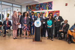 Gruppe von Menschen vor einem Podium mit einem Mikrofon und einer Tafel mit der Aufschrift "Sancho Stadtverordneter tritt dem Stadtrat bei", mit sitzenden und stehenden G├Ąsten, Fahnen, Feuerl├Âscher und Deckenleuchten im Hintergrund.