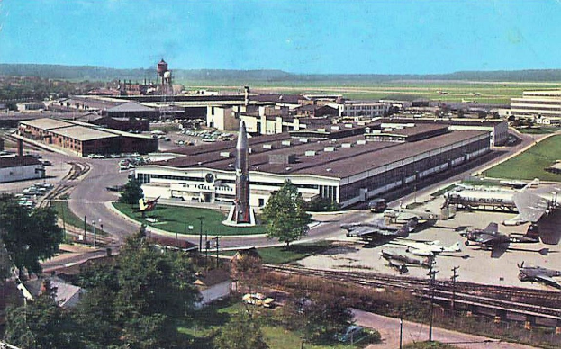 Alte Schwarz-Weiß-Flughafenszene mit parkenden Flugzeugen, Gebäuden, Bäumen, Straßen, Fahrzeugen, Strommasten, Gras, Bahnschienen, fernen Türmen, Hügeln und einem klaren Himmel; Text erscheint entlang der unteren Kante.