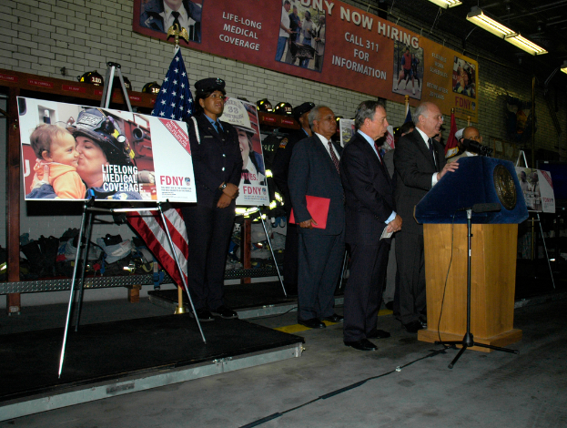 Eine Gruppe von Menschen in der Nähe eines Podiums mit Mikrofonen, umgeben von Schildern auf Ständern, Fahnen und Schildern an der Wand, alles unter hellen Lichtern.