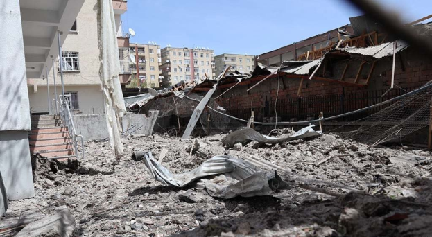 Die Nachwirkungen eines israelischen Luftangriffs in Gaza zeigen ein zerstörtes Gebäude, das zu Schutt und Asche geworden ist, auf einer Straße mit eingestürzten Wänden, Metallstangen, einem Zaun, einer Treppe, benachbarten Gebäuden mit Fenstern, Bäumen und einem bewölkten Himmel.