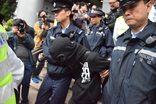 Eine Gruppe von Polizeibeamten steht vor einer Menge, einige tragen Mützen und halten Kameras, mit einem Gebäude mit Säulen und Deckenleuchten im Hintergrund und Bäumen links.