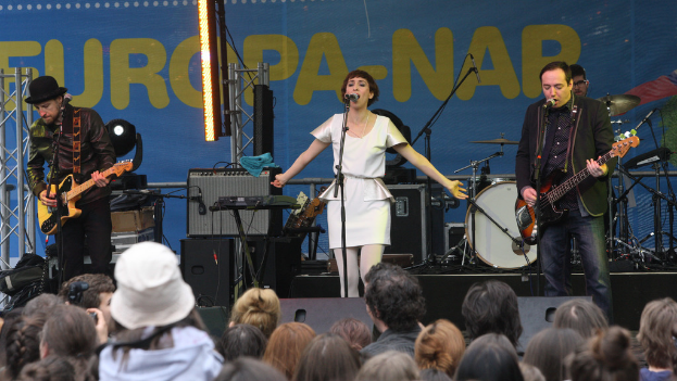 Eine Bühne mit vier Musikern: Eine singende Frau in weißem Kleid in der Mitte, zwei Männer mit Gitarren auf der linken und rechten Seite und eine Menge und Lautsprecher im Hintergrund.