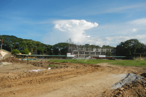 Ein Schotterweg, der zu einem teilweise errichteten Gebäude führt, umgeben von Gras, Strommasten und Bäumen unter einem bewölkten Himmel.