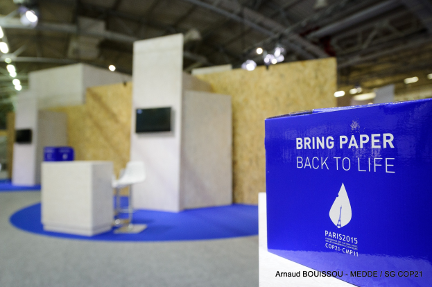 Messestände mit blauer und weißer "Bring Paper Back to Life"-Aufschrift auf der rechten Seite, mit unscharfen Stühlen, Tischen, einem wandmontierten Fernseher und Deckenleuchten im Hintergrund.