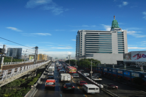 Eine städtische Straße mit starkem Verkehr, hohen Gebäuden, einer Brücke auf der linken Seite, Strommasten mit Drähten, Bäumen und einem bewölkten Himmel im Hintergrund.