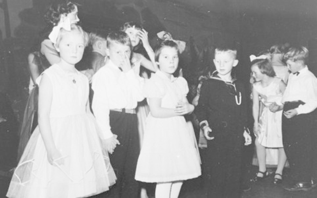 Schwarzes und weiße Bild einer Gruppe von Kindern, die auf einem Tanzboden stehen und lächeln, mit einer Wand im Hintergrund.