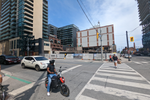 Ein Mann fährt auf einem Fahrrad eine belebte Straße mit hohen Gebäuden entlang, trägt ein schwarzes T-Shirt, blaue Jeans und einen schwarzen Helm, mit Menschen, Autos und Verkehrszeichen drumherum.