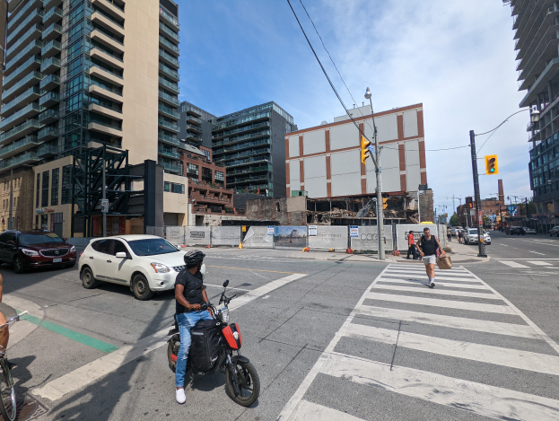 Ein Mann fährt auf einem Fahrrad eine belebte Straße mit hohen Gebäuden entlang, trägt ein schwarzes T-Shirt, blaue Jeans und einen schwarzen Helm, mit Menschen, Autos und Verkehrszeichen drumherum.