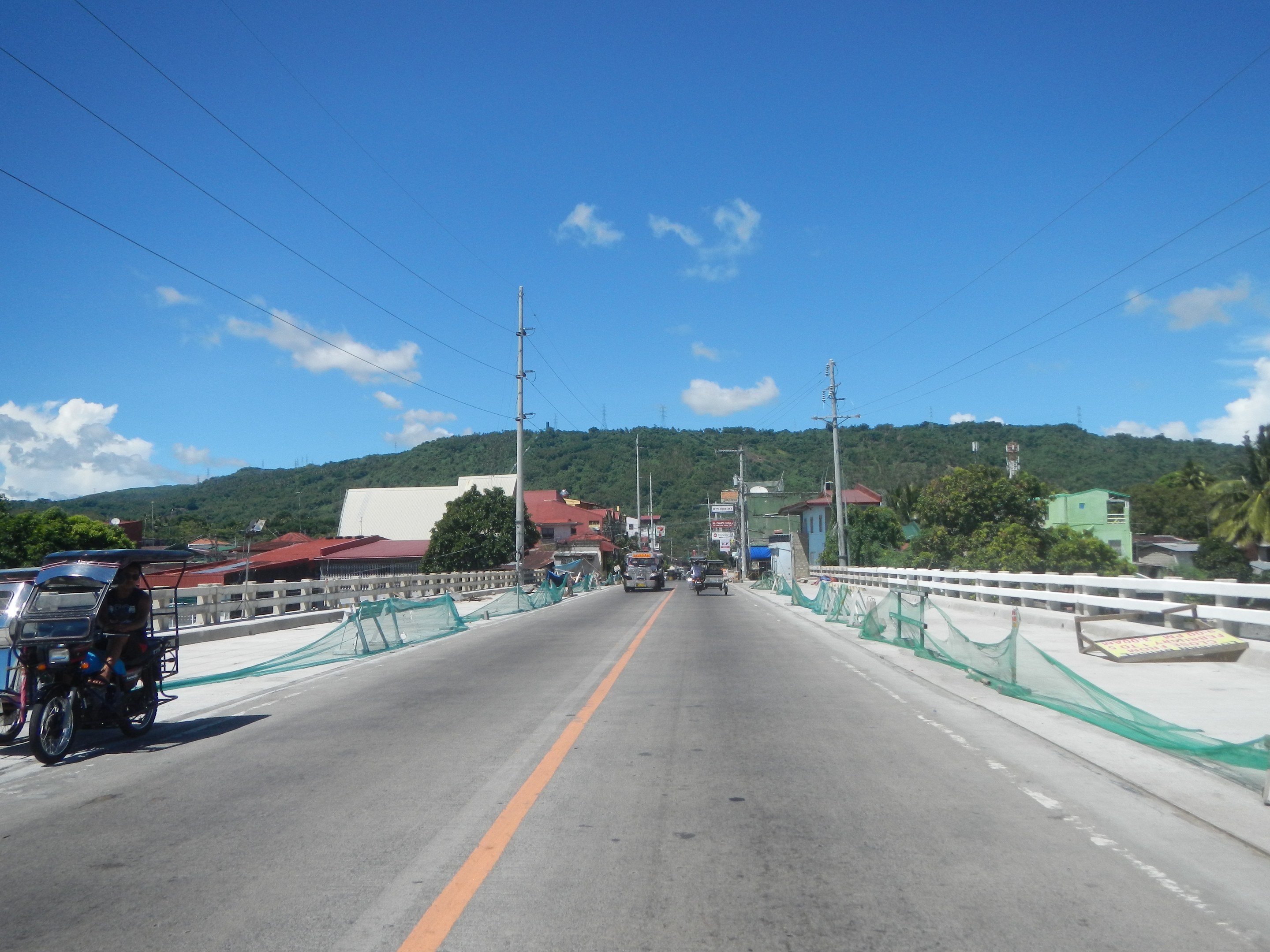 Autobahn mit fahrenden Fahrzeugen, ein geparktes Motorrad auf dem Seitenstreifen, Geländer, Strommasten mit Drähten, Bäume, Gebäude, entfernte Hügel und ein bewölkter Himmel.