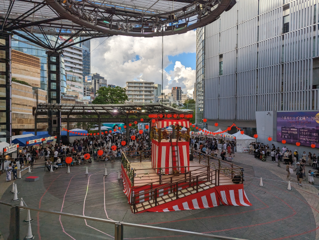 Große Menschenmenge versammelt sich um eine Bühne in einer städtischen Umgebung mit Gebäuden, Bäumen und einem bewölkten Himmel, mit Künstlern und Equipment für die Eröffnung des Tokyo Circus Festivals.