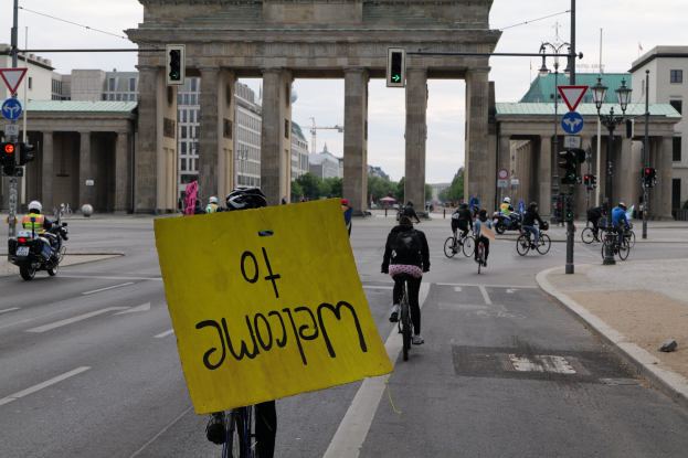 Eine Gruppe von Radfahrern fährt am Brandenburger Tor in Berlin vorbei, einer hält ein gelbes Schild, mit Laternenmasten, Verkehrsampeln, Gebäuden und Bäumen im Hintergrund unter einem klaren Himmel.