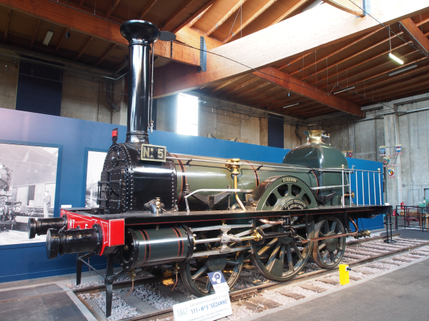 Alte Dampflokomotive in einem Museum ausgestellt, auf Schienen positioniert mit Informationsschildern und Ständen in der Nähe, vor einer Wand mit einem gerahmten Foto und Deckenbeleuchtung.