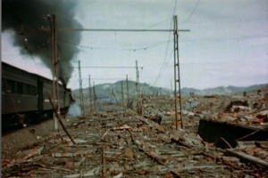 Ein Zug fährt durch ein beschädigtes Gebiet mit Rauch, der aus ihm aufsteigt, umgeben von Strommasten und Drähten, mit Hügeln und einem klaren Himmel im Hintergrund und verstreutem Schutt auf dem Boden.