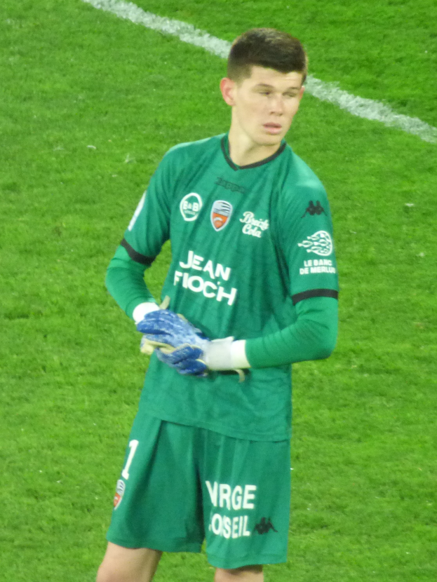 Ein Mann in grüner Torwarthose, grünem T-Shirt und blauen Handschuhen steht auf einem Fußballfeld.