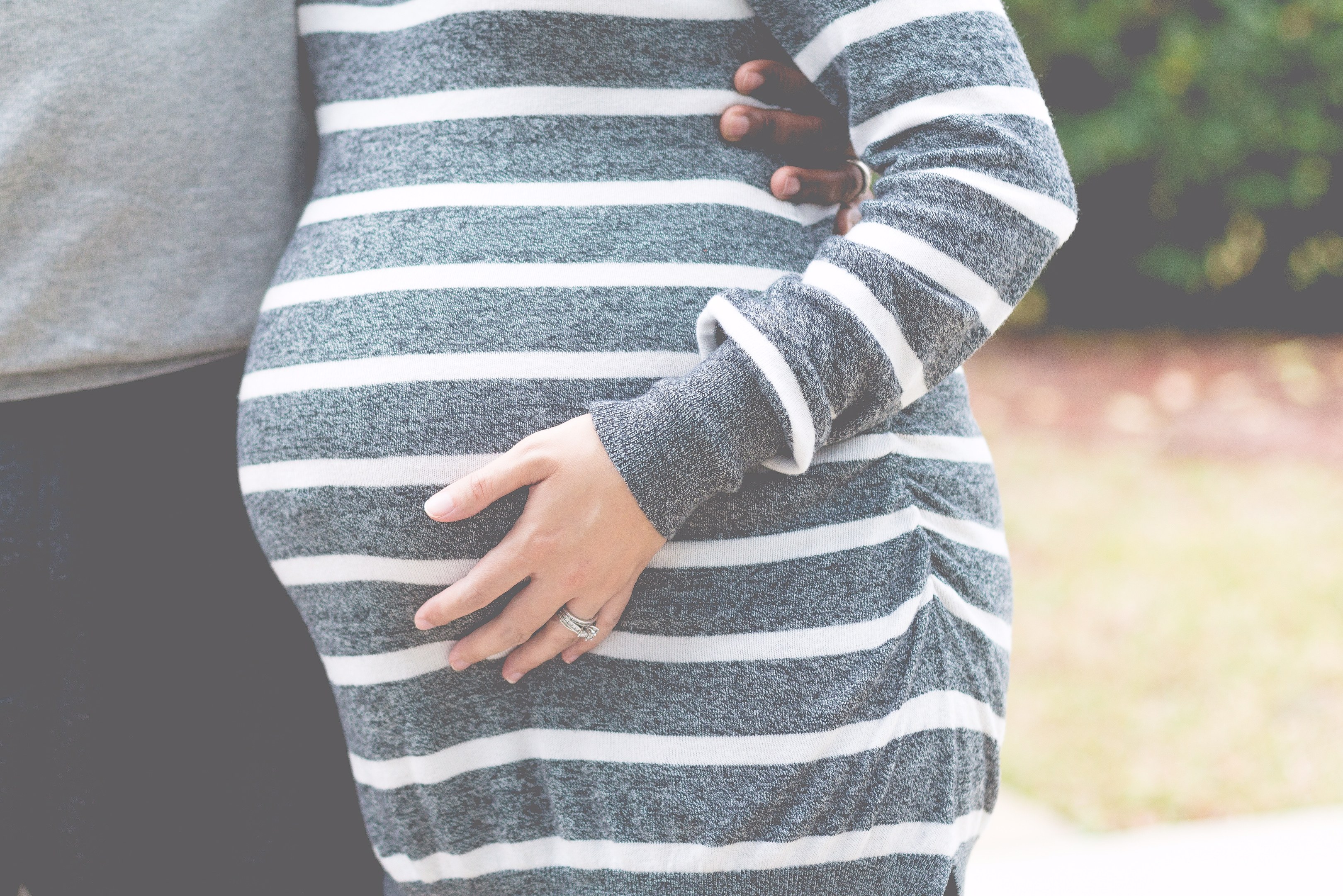 Schwangere Paare nebeneinanderstehend mit Händen auf dem Bauch, tragend Maternity-Kleidung, in einer holzigen Outdoor-Umgebung.