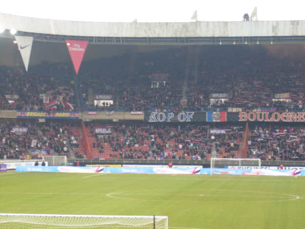Ein grasbewachsener Stadionboden mit Torpfosten auf beiden Seiten, umgeben von sitzenden Zuschauern und geschmückt mit zahlreichen Werbetafeln, unter einem klaren Himmel.