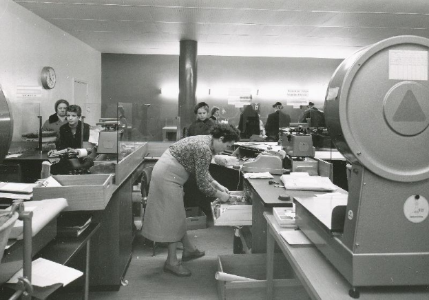 Schwarzes und weißes Büro-Szenario mit Menschen an Schreibtischen, einige stehend und einige sitzend, Papiere und Gegenstände auf Tischen, eine Wanduhr, eine Säule und sichtbare Decke.