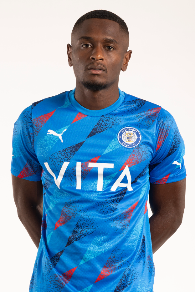 Ein Mann in einem blauen Leicester City Away-Shirt von der Saison 2018-19, der entspannt mit händen in den Taschen und einem leichten Lächeln dasteht.