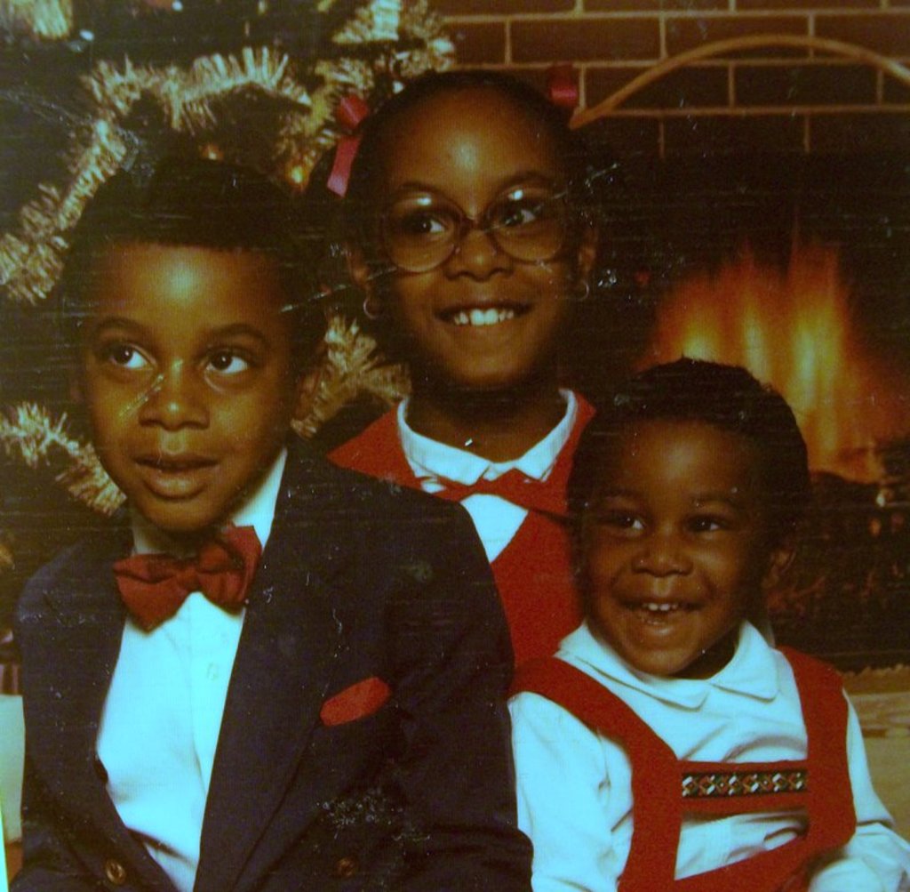 Drei Kinder lachen vor einem Weihnachtsbaum mit einem Kamin und einem Holzstapel im Hintergrund.