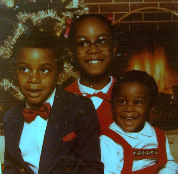 Drei Kinder lachen vor einem Weihnachtsbaum mit einem Kamin und einem Holzstapel im Hintergrund.