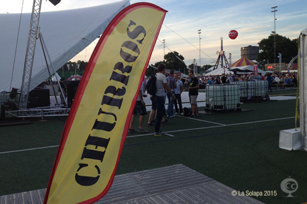 Eine KarnevalsSzene mit Zelten, einem zentralen Mast, Menschen, Bäumen, Strommasten, Straßenbahnen auf der Straße und einem eisernen Stand auf der linken Seite unter einem klaren Himmel.