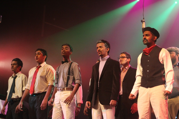 Eine Gruppe von M├Ąnnern auf einer B├╝hne, die ein Drama auff├╝hren, mit gr├╝nen Spotlights im Hintergrund.