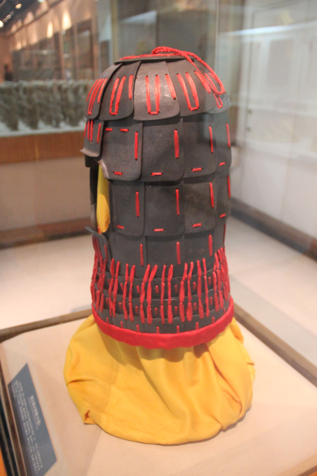 Ein Helm, der in einer Glasvitrine mit einem gelben Tuch darüber ausgestellt wird, begleitet von einer Texttafel links und Deckenlampen im Hintergrund.