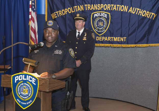 Zwei Polizeibeamte stehen an einem Rednerpult mit einem Mikrofon; einer hält Papiere, während im Hintergrund ein Banner, eine Flagge und ein Tisch zu sehen sind.