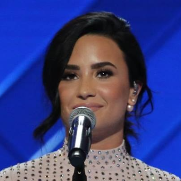 Demi Lovato steht mit einem Mikrofon auf der Bühne bei den Grammys, trägt ein schwarzes Kleid mit einem eleganten Bob, vor einem blauen Hintergrund, mit einem selbstbewussten Gesichtsausdruck.