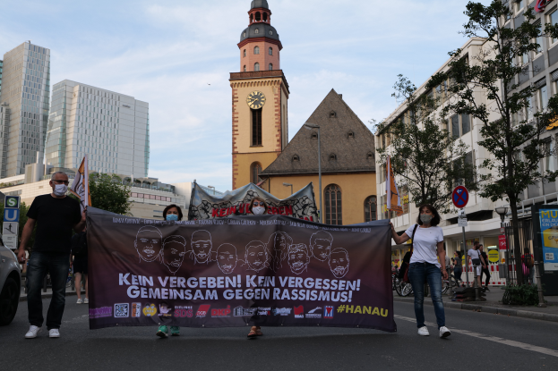Eine Gruppe maskierter Menschen geht eine Straße entlang und hält eine Protestfahne, mit einem geparkten Auto auf der linken Seite, Gebäuden, Bäumen, Schildern, Polen und einem Uhrenturm im Hintergrund unter einem klaren blauen Himmel.
