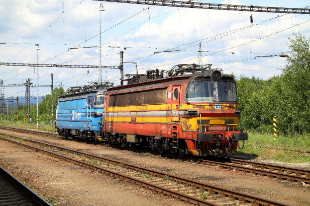 Eine Zugmaschine auf Schienen mit Strommasten, Kabeln, Bäumen, Pflanzen und Wolken am Himmel.