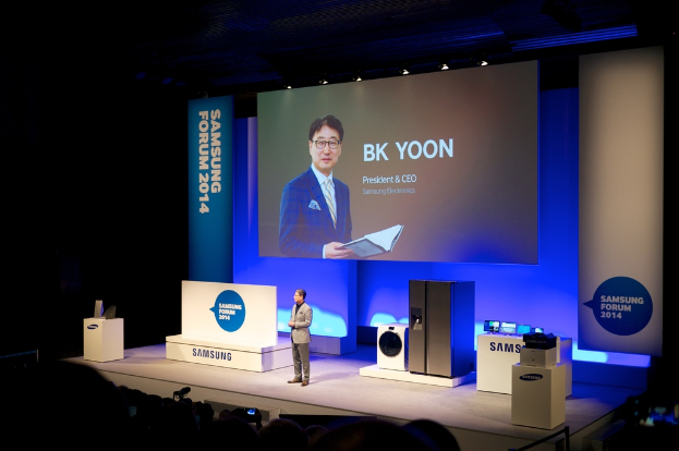 Ein Mann steht vor einer Whiteboard und einem kühlschrankähnlichen Objekt, hält ein Buch und lächelt, mit Text neben ihm, während Menschen auf Stühlen in einem Konzertsaal mit beleuchteter Decke sitzen.